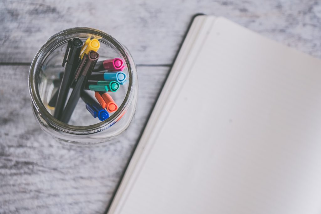 Jar with coloured pen on a table next to an open notebook turned to a blank page