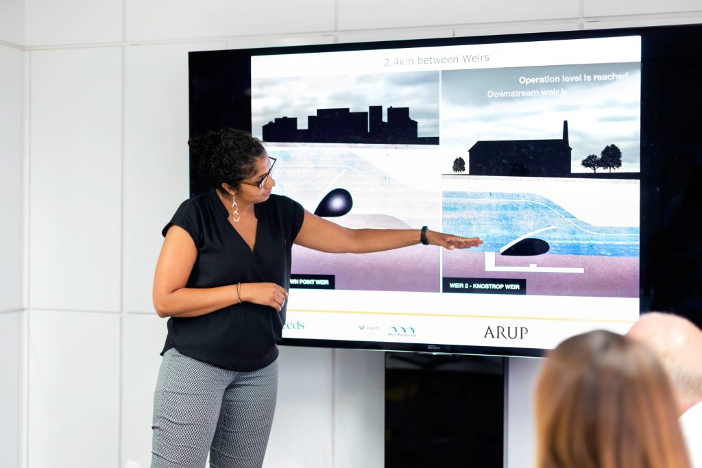 A person standing at the front of the room pointing to a presentation on screen.